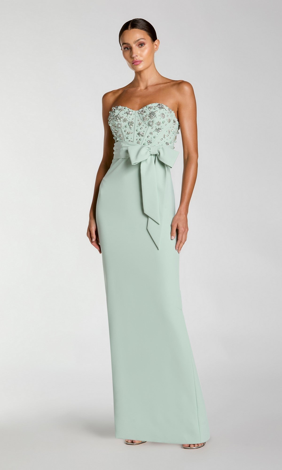 A woman wearing the Mac Duggal 2395 long formal dress in sage, featuring a strapless design with floral embellishments on the bodice and a statement waist bow, stands against a plain background.
